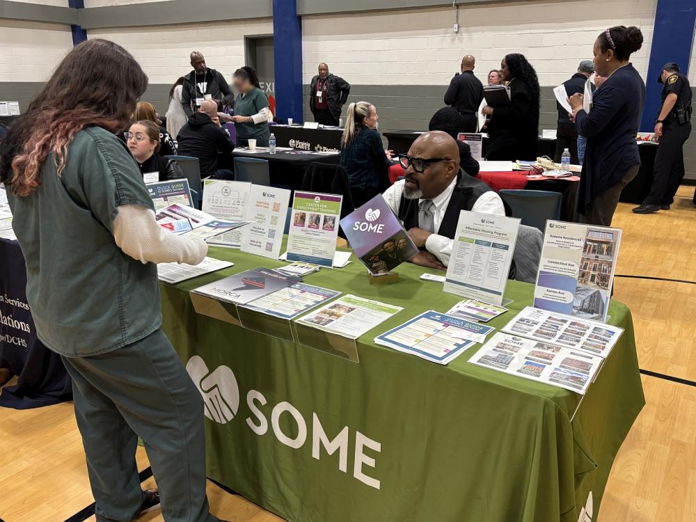 Inmate in a green jumpsuit speaking with an exhibitor who has printed materials at his table