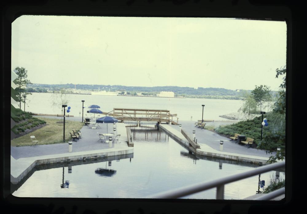 Photo showing a restored park with a walking bridge over a filled pool in the shape of the canal, with the Potomac River in the background.