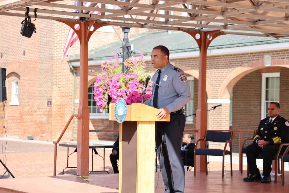 A police officer in uniform speaking at a podium with a floral arrangement, under a pergola in a public outdoor setting.