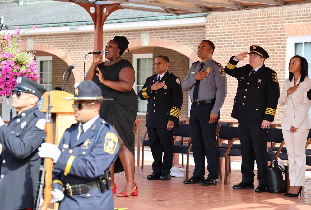 A person sings during an outdoor event while several uniformed police officers, including some in formal attire with medals, stand at attention saluting.