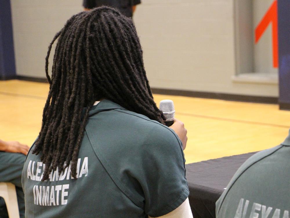 Seen from behind, an inmate seated and wearing green jumpsuit that says Alexandria Inmate is holding a microphone in front of his unseen face