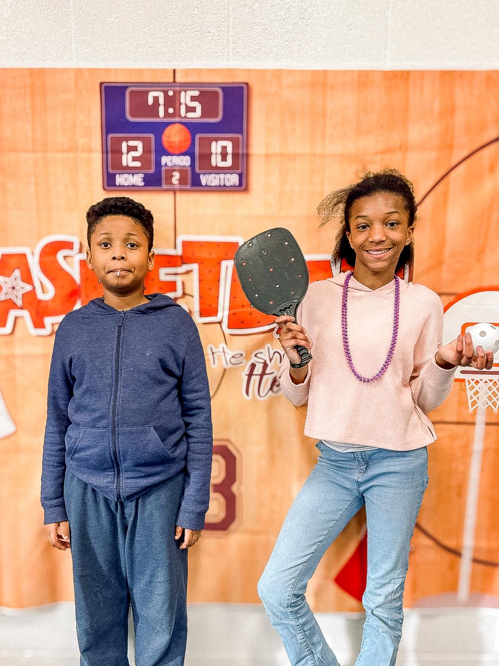 Teenage boy standing next to teenage girl who's holding a pickleball
