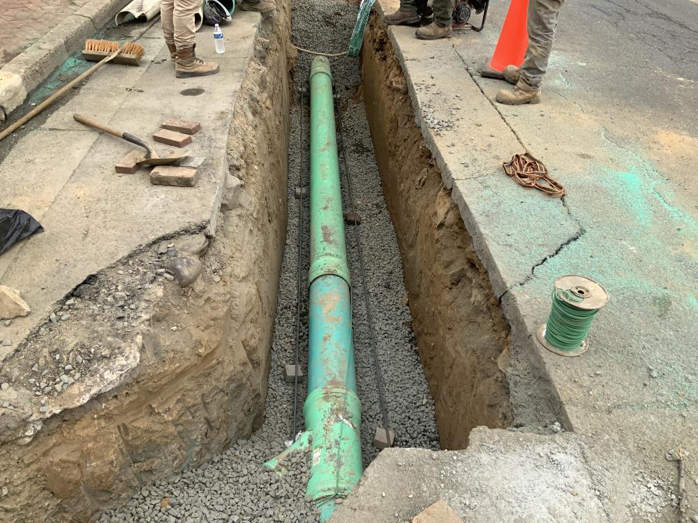 A turquoise sewer pipe is set inside a gravel-lined trench cut into the pavement of the road.