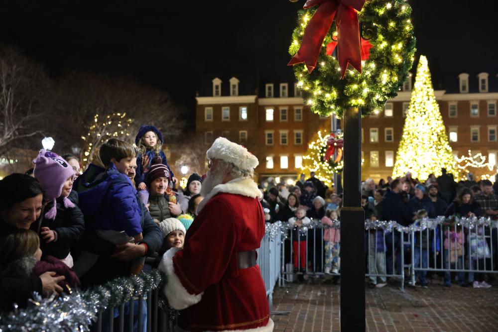 A man dressed as Santa greets a young child. A lighted holiday tree and City Hall can be seen in the background.