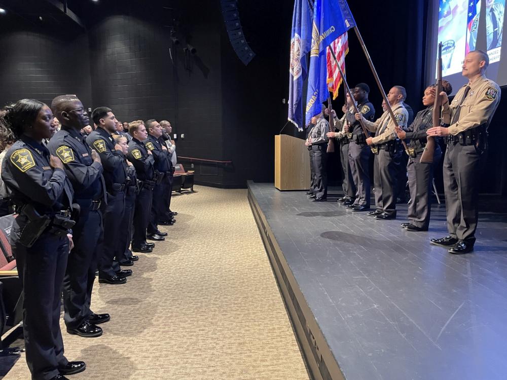 Honor guard of deputies in ceremonial uniforms presenting the colors while audience stands and salutes flag