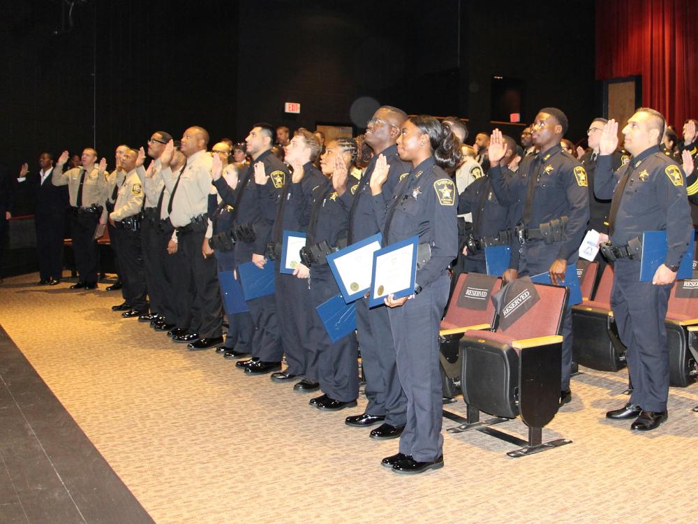 Two rows of new deputies, some in blue uniforms and some in tan, holding up their right hands and taking an oath at a ceremon