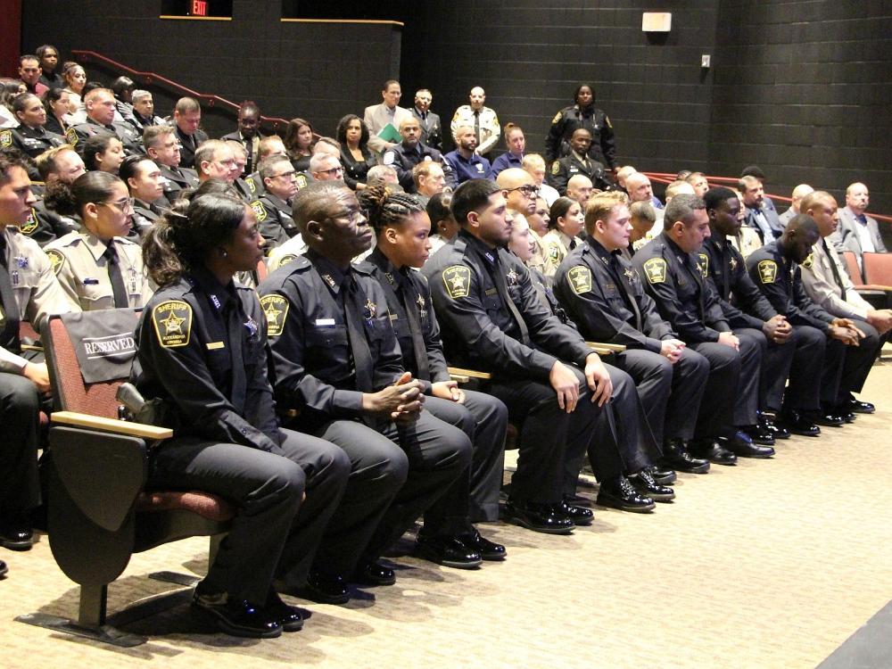 Rows of recruit deputies, some in blue uniforms and some in tan, seated at their graduation ceremony