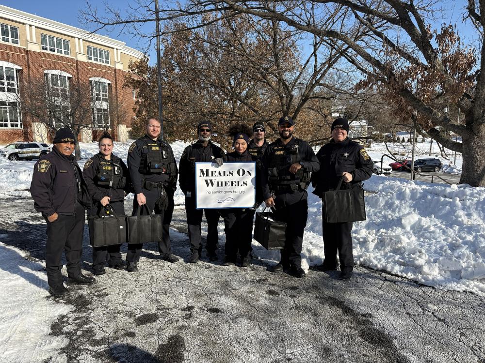several deputies in blue winter uniforms holding sign that says Meals on Wheels with lots of snow in the background