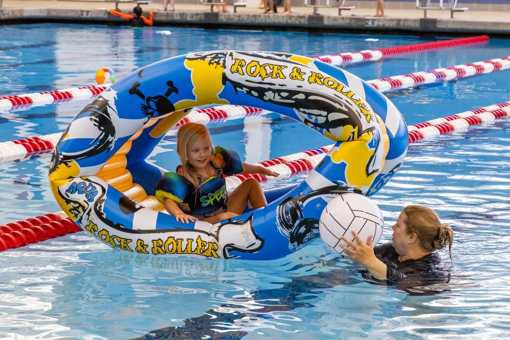 A child sitting inside one blow up pool toy while her mother, who's standing next to her, hands her a blow up volleyball pool toy