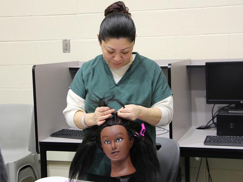 inmate in green uniform braiding the hair of a mannequin head