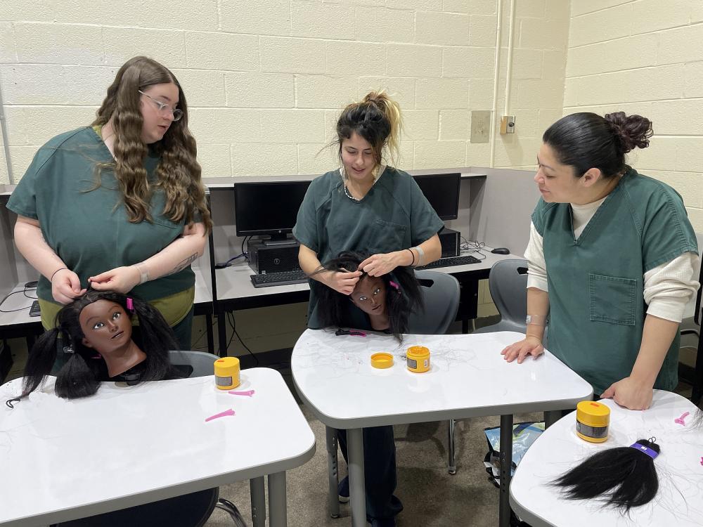 Three inmates in green uniforms in a hair braiding class, with two of the students braiding the hair of mannequin heads