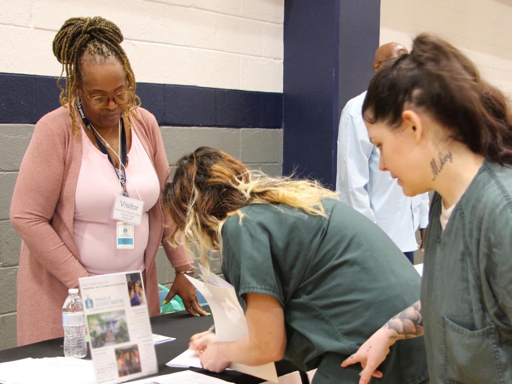 Two inmates in green uniforms looking at information while a member of Friends of Guest House assists them at the Reentry Resource Fair