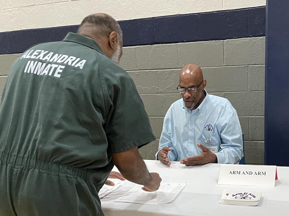 Inmate in green uniform that says Alexandria Inmate on the back speaking with a member of Arm & Arm peer support group at Reentry Resource Fair