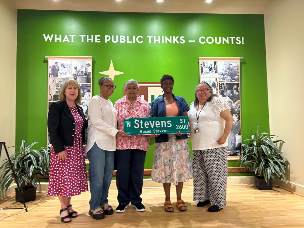 A group of five people standing in front of a display with the text "WHAT THE PUBLIC THINKS – COUNTS!" They are holding a green street sign that reads "N Stevens - Moses Stevens St." The background includes historical photos, and there are plants on both sides.