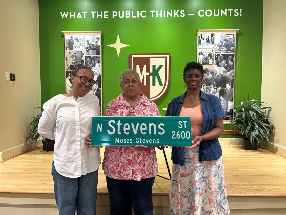 Three people stand on a wooden stage holding a "N Stevens St" street sign with "Moses Stevens" written underneath. Behind them is a display featuring historical photos, with a wall sign that reads "What the Public Thinks - Counts!" and a logo with the letters "MK".