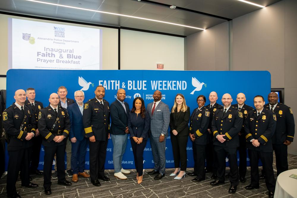 Uniformed APD personnel and guests pose for a group photo in front of a blue Faith & Blue Weekend backdrop at the inaugural Prayer Breakfast.