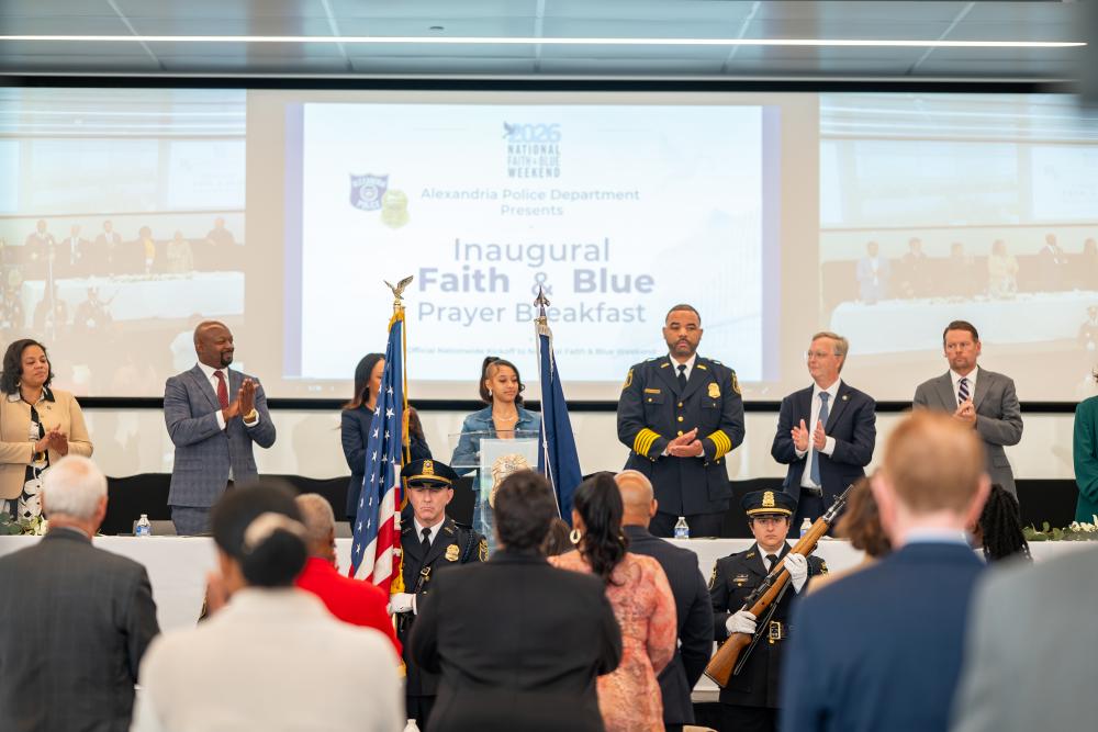 APD honor guard members present flags as attendees stand and guests at the head table applaud during the inaugural Faith & Blue Prayer Breakfast. A screen behind them displays the event title.