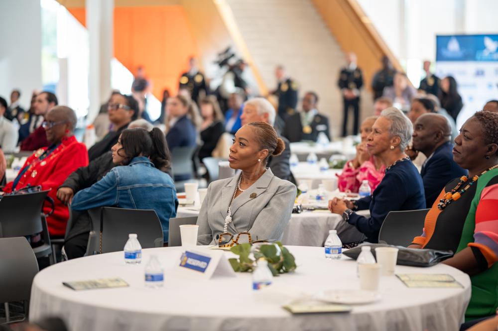 Attendees seated at round tables listen to the program during APD’s inaugural Faith & Blue Prayer Breakfast, with uniformed officers standing in the background.