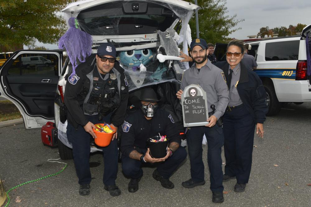 Four adults pose beside an open vehicle hatch decorated with large cartoon eyes, pointed teeth, cobwebs and hanging fabric.
