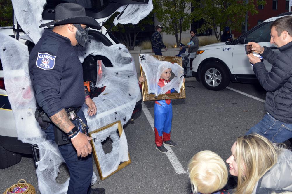 An officer wearing a black hat and skull face covering stands beside a cobweb-covered vehicle as an adult holds up a phone to take a photo.