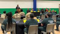sheriff speaking at podium with several people seated and applauding inmates who are seated and wearing tan and green jumpsuits