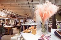 In the foreground is a round display rack with several colorful shoes on display around a glass vase of pale pink pampas grass. In the background are racks of clothing on white hangers, some on wheels, and displays decorated with string lights.