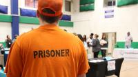 View from behind an inmate in an orange baseball cap and orange shirt with the world 'prisoner' on the back, as the inmate looks toward tables and people at a resource fair