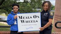 teen worker and deputy in uniform holding a sign that says Meals on Wheels