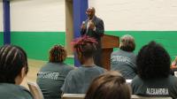 Man in a suit holding a microphone and speaking to several inmates who are seated at tables, wearing green jumpsuits and only visible from behind.