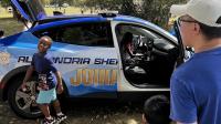 Child smiling outside blue sheriff's cruiser with adult and child looking on