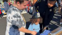 Civilian and deputy in blue uniform assisting a small child in getting fingerprinted at a community event.