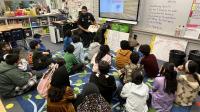 Deputy in blue uniform seated and reading a book to school children sitting on the floor of a classroom.