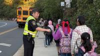 Several school children walking down a sidewalk away from the camera and Sheriff in blue uniform and yellow safety vest exchanging fist bump with one of the kids. A school bus is visible in th background.