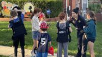 Deputy talking with several children outside a home with two kids trying on black vests, including one that says "Sheriff"