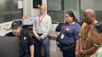 Deputy in blue uniform seated and looking at computer screens while four civilians look on and listen