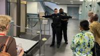 Inside a couthouse, two deputies in blue uniforms demonstrating how to scan and search someone after passing through a magnatometer while five citizens look on