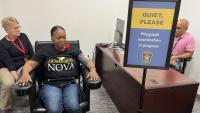 Two women sitting in chairs, one of them with cords attached to her hands and body, with an investigator seated at a desk looking at a computer screen, and a sign reading Quiet please, polygraph examination in progress