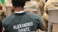 Inmate in a green jumpsuit, seated and only visible from behind, holding his hands up as he listens to music, with other inmates wearing tan jumpsuits seated nearby