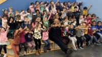 Deputy in a blue uniform with about 30 small children around him inside a school, all giving thumbs up