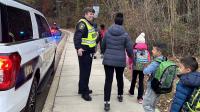 Sheriff in blue uniform and bright yellow traffic vest watching as several small children and an adult walk to school down a sidewalk. A white law enforcement SUV with its lights on is parked to the left.