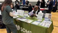 Inmate in a green jumpsuit speaking with an exhibitor who has printed materials at his table