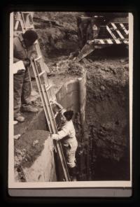 An archaeologist climbs a ladder that leans against the inset tide lock gate stone wall. 