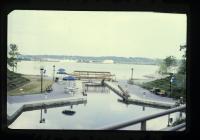 Photo showing a restored park with a walking bridge over a filled pool in the shape of the canal, with the Potomac River in the background.