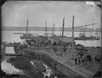 Black and white photo of the coal wharf at the canal's tide lock. Tall ships are shown in the Potomac River in the background. The foreground shows the canal lock and workers at the wharf near piles of coal.