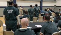 Several inmates in green jumpsuits, some standing and seated, in a gymnasium visible from behind