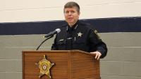 Sheriff in dark blue uniform standing at podium when the Sheriff's Office star on it and speaking into a microphone