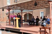 An officer speaks at a podium adorned with flowers at an outdoor event at Market Square, with seated attendees in uniforms and formal attire listening attentively.