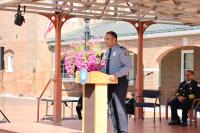 A police officer in uniform speaking at a podium with a floral arrangement, under a pergola in a public outdoor setting.