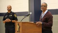 Deputy in a blue uniform standing next to a civilan employee who is speaking at a podium