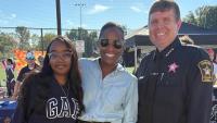 Sheriff in blue uniform wearing a pink badge and standing next two community members at an outdoor event